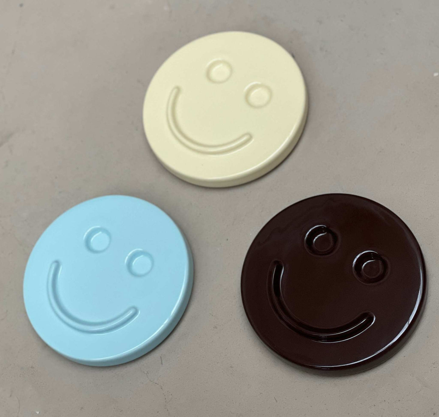 Three ceramic smiley face artworks in blue, cream, and brown, handcrafted by Edith Beurskens.